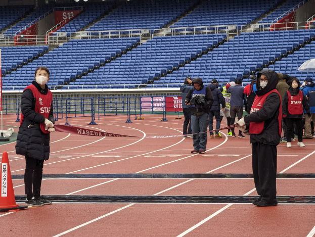 Gruppe von Menschen an der Startlinie eines Rennens, die ein Band halten, mit einem Verkehrskegel links und Event-Aufbau im Hintergrund.