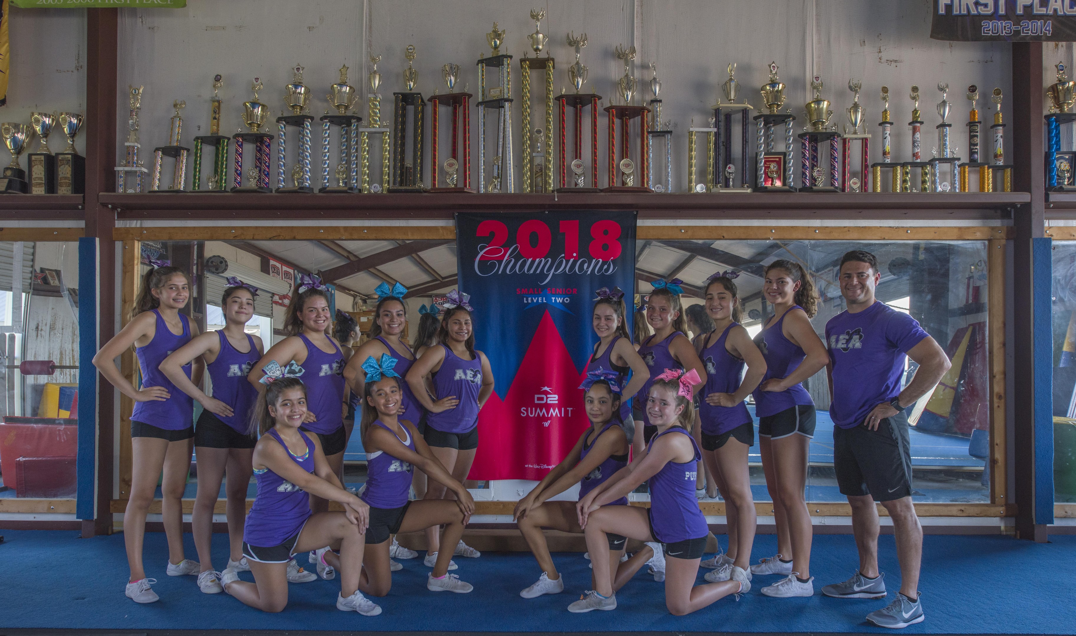 Gruppe von Cheerleadern in lila T-Shirts und schwarzen Shorts, die vor einem Vitrinenschrank mit einer '2018 Summit Cheerleading Champions'-Banner im Hintergrund posieren.