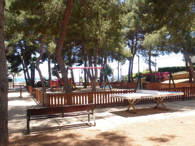 Eine Parkszene mit einem zentralen Tischtennistisch, umgeben von Bänken, einem Zaun, Schaukeln, Pfosten, Pflanzen und Bäumen, mit einem Wasserlauf und einem klaren blauen Himmel im Hintergrund.