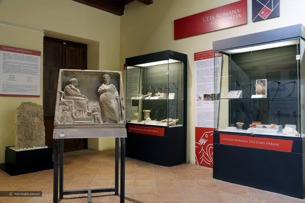 Ein Museumsraum in Rom mit verschiedenen Artefakten, darunter Skulpturen auf Ständern, Glasvitrinen mit Objekten, Informationsschilder an den Wänden und eine Tür, mit einem Wasserzeichen unten.