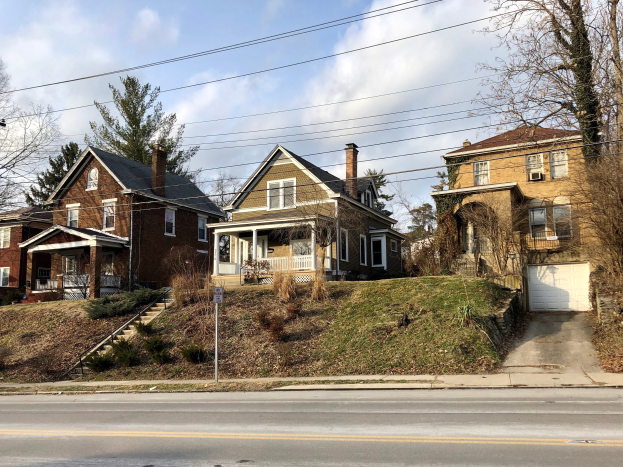 Reihe von Häusern mit Fenstern, Türen, Säulen, Geländern, Stufen, Pflanzen, Gras und einem Pfahl mit einer Tafel, vor einem bewölkten Himmel.