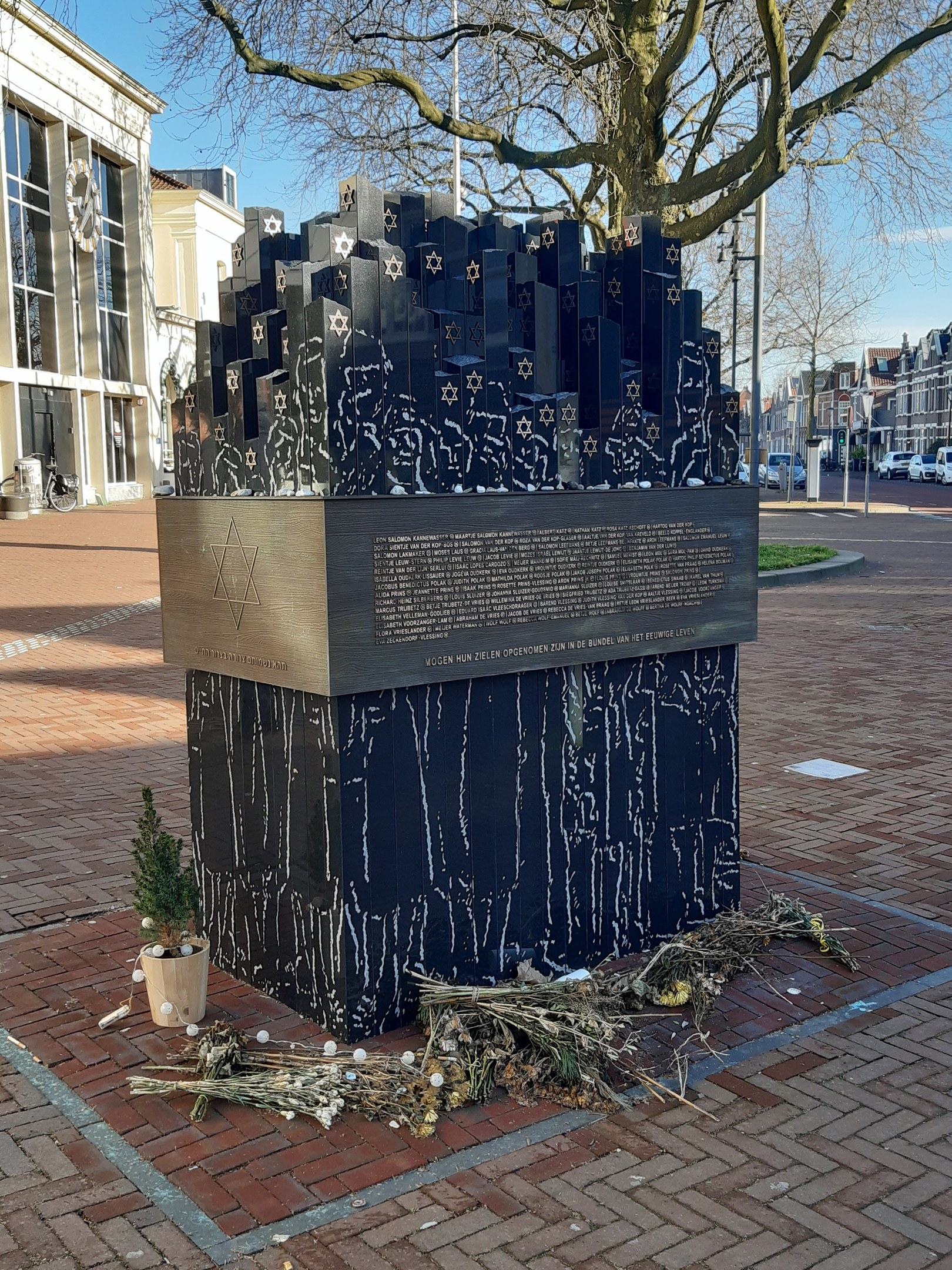 Denkmal für Holocaust-Opfer in einem Stadtplatz, mit einem Baum davor, einem Topf mit einer Pflanze, verstreuten trockenen Blättern und Gebäuden im Hintergrund.