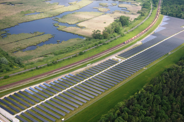 Luftaufnahme einer Solarpark mit Solarmodulen, umgeben von Bäumen, Gras, Pflanzen und Wasser, mit einem Zug auf einem Bahngleis.