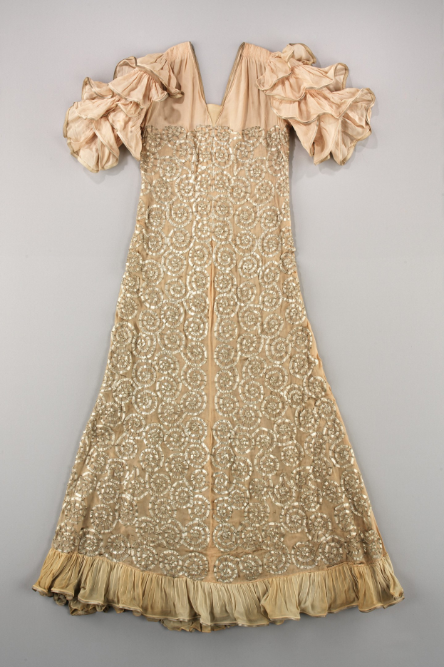 Ein cremefarbenes Kleid mit goldener Stickerei und Rüschenärmeln, das auf einer weißen Oberfläche präsentiert wird und mit detaillierten Mustern und Designs verziert ist.