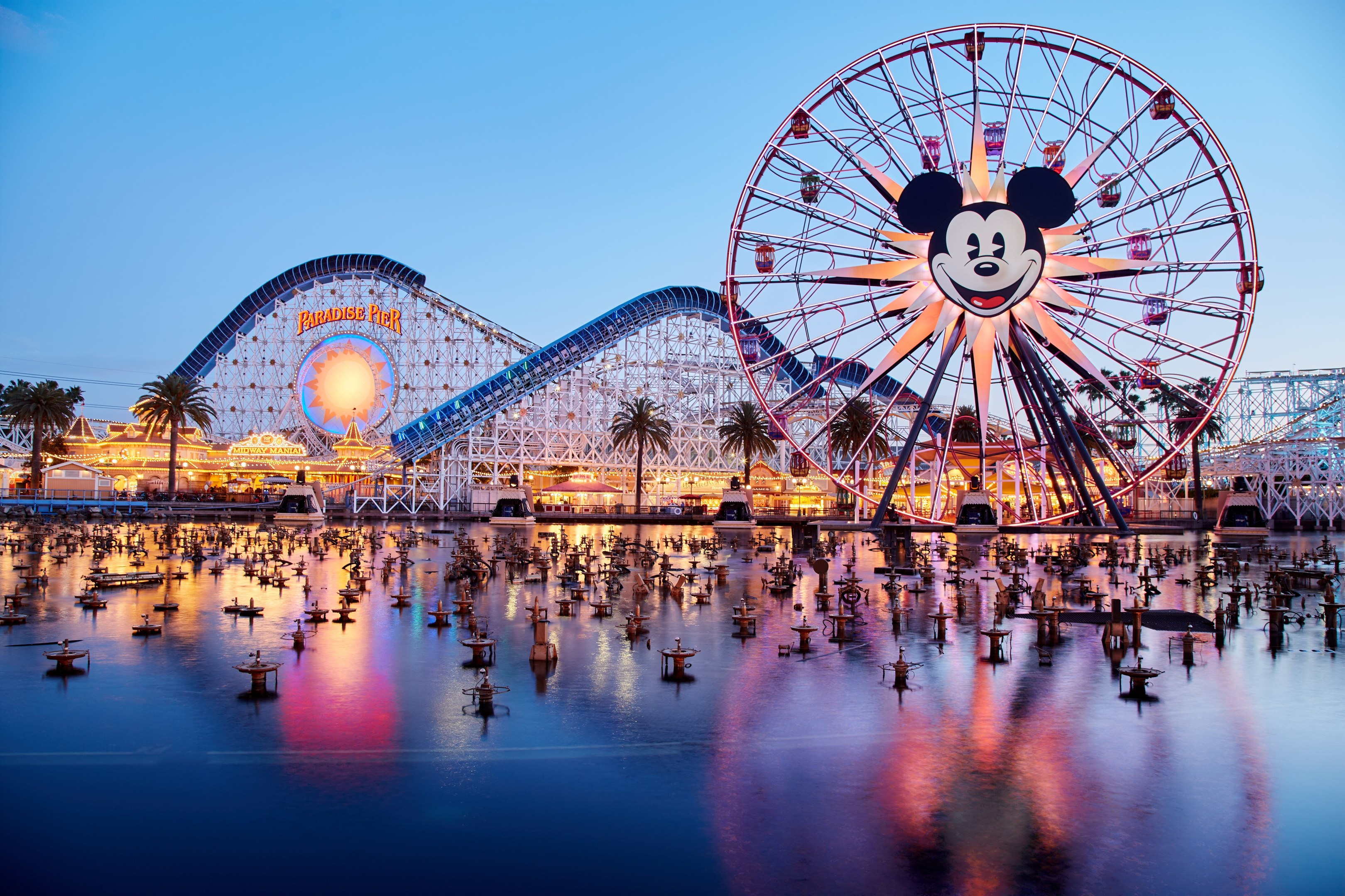 Eine lebendige Dämmerungsszene im Disneyland California Adventure mit einem riesigen Mickey-Maus-Rad im Vordergrund, einer Achterbahn im Hintergrund und Booten auf dem Wasser, mit einem bunten Himmel und Silhouetten von Bäumen.