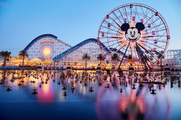 Eine lebendige Dämmerungsszene im Disneyland California Adventure mit einem riesigen Mickey-Maus-Rad im Vordergrund, einer Achterbahn im Hintergrund und Booten auf dem Wasser, mit einem bunten Himmel und Silhouetten von Bäumen.