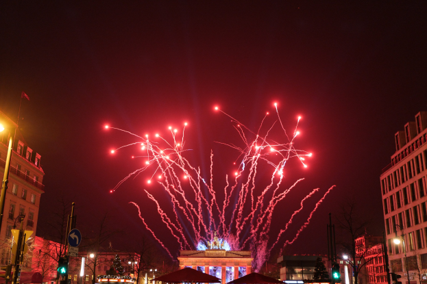 Eine nächtliche Straßenansicht in Berlin an Silvester, mit Gebäuden, Bäumen, Laternen, Ampeln, Schildern, Zelten, Menschen und einem Feuerwerk am Himmel.