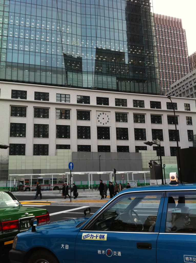 Eine Szene mit mehreren Autos, Holzplanken, einem Bus, Pfählen, Gebäuden und einigen Menschen, die stehen und Text auf einem der Autos zu erkennen ist.