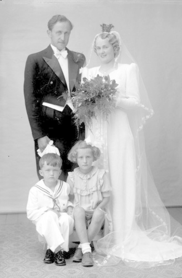 Schwarzes und weißes Foto einer Braut in einem weißen Kleid, die einen Strauß hält, ein strahlender Bräutigam neben ihr und zwei Kinder, die vor ihnen auf dem Boden sitzen.