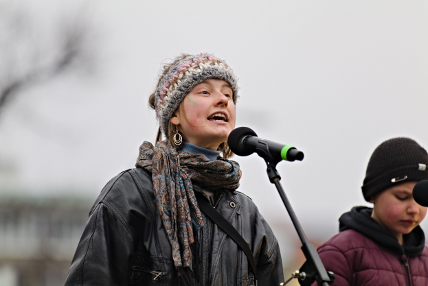 Eine Frau in einer schwarzen Jacke, einem Schal und einer Mütze singt in ein Mikrofon auf der Bühne, während eine weitere Person in einer maronenfarbenen Jacke und einer schwarzen Mütze neben ihr steht, und ein unscharfer Hintergrund bildet.