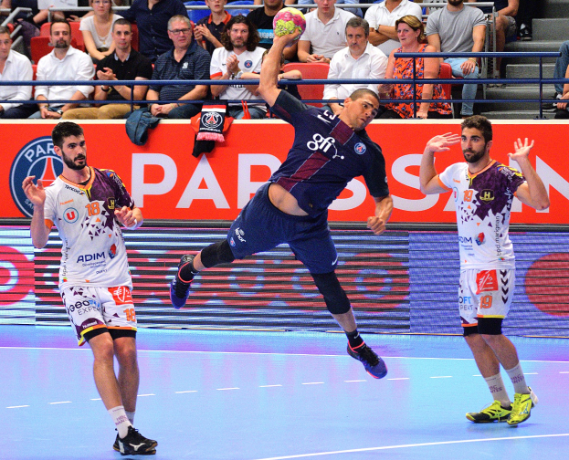 Mannergruppe spielt Handball auf einem Platz mit einem Ball im Spiel, Zuschauer im Hintergrund und verstreute Kleidung in der Nähe des Platzes.