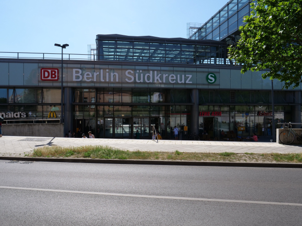 Ein große, gläserne Bahnhofsgebäude mit der Aufschrift "Südkreuz" in Berlin, Deutschland, mit Straßenmasten, Lichtern, Fahrzeugen, Füpfen, Fahrrädern, Bäumen und einem klaren blauen Himmel.