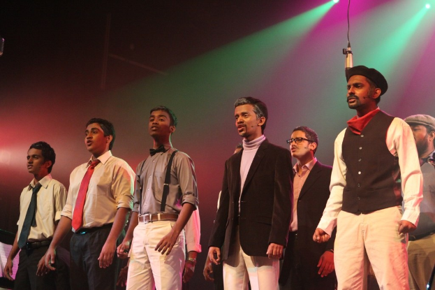 Eine Gruppe von Männern führt ein Drama auf der Bühne auf, während grüne Scheinwerfer im Hintergrund leuchten.