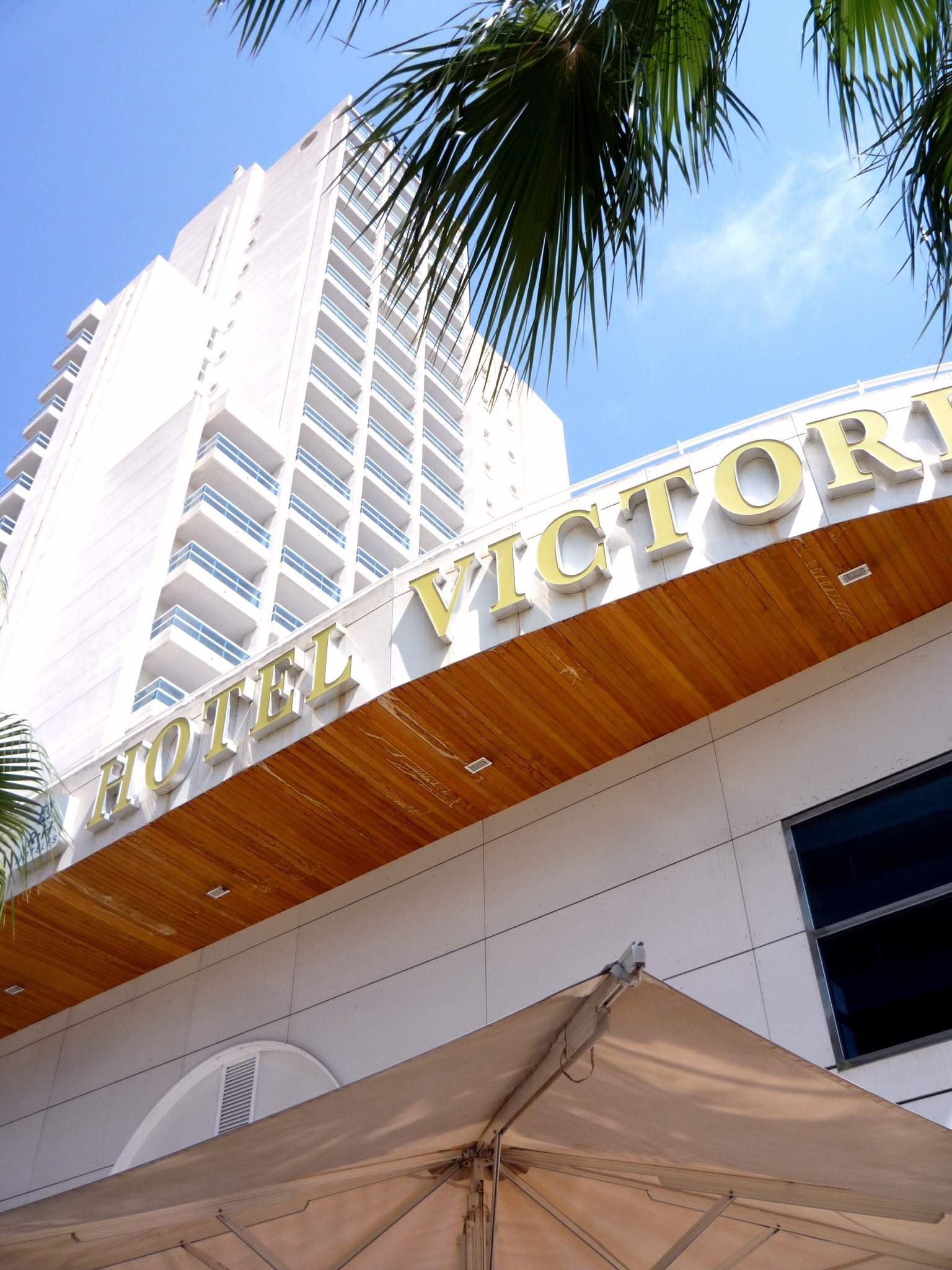 Das Bild zeigt das Hotel Victoria in Miami Beach, Florida, einen hohen Turm mit Glasfenstern und einem Namen auf der Wand, mit einem Zelt und einem Baum unten und weißen, flauschigen Wolken am Himmel.