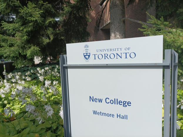 Das Bild zeigt die Außenseite des neuen Colleges der University of Toronto am Wetmore Hall, mit einem Schild davor, umgeben von grünem Gras, bunten Blumen, hohen Bäumen und einem hellblauen Himmel.