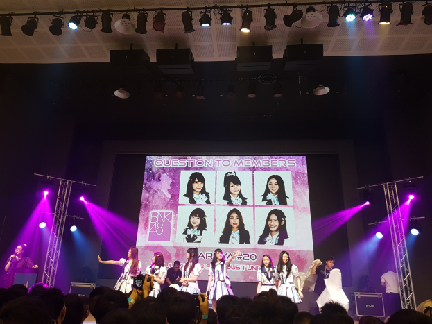 Eine Gruppe von Menschen tritt auf einer hell beleuchteten Bühne mit Tr├Ągern auf, w├Ąhrend eine Menge darunter das Ereignis aufzeichnet; ein gro├če Bildschirm im Hintergrund zeigt Bilder der K-Pop-Gruppe BK48.