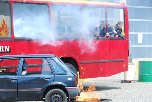 Ein roter Doppeldeckerbus mit Rauch, daneben ein parkendes Auto, drei sichtbare Passagiere und im Hintergrund ein Gebäude mit Glasfenstern und einem Fass.