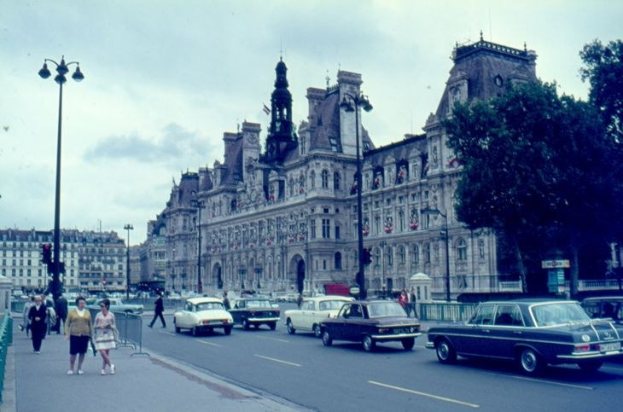 Ein altes Foto einer Stadtstraße mit Autos, Fußgängern, Straßenlaternen, Geländern, Bäumen, Gebäuden mit Fenstern und einem bewölkten Himmel.