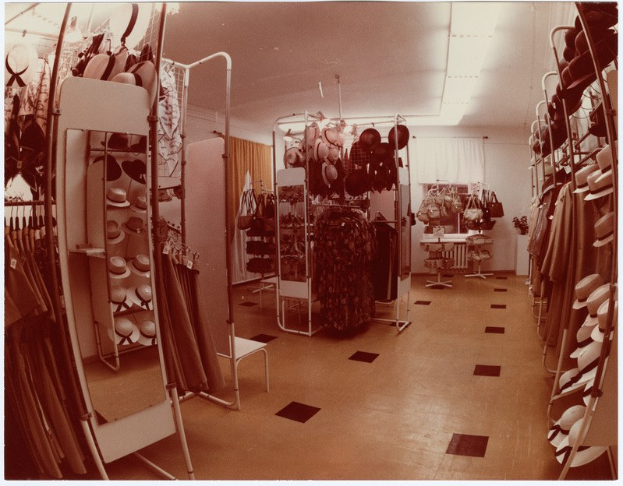 Altes Foto eines Kleiderladen-Interieurs mit Kleiderstangen, Hutständer, Spiegel, Stuhl, Tisch mit Gegenständen, Vorhängen und Deckenleuchten.