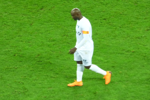 Ein Fussballspieler in einem weissen T-Shirt, weissen Shorts und orangefarbenen Schuhen läuft über ein saftiges grünes Feld.