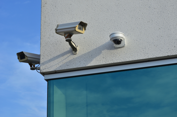 Zwei Überwachungskameras, die an der Seite eines Gebäudes montiert sind, mit einer Glaswand im Vordergrund und einem klaren blauen Himmel im Hintergrund.