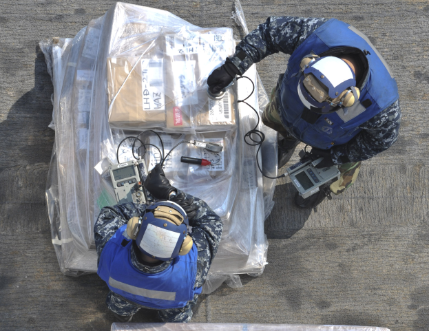 Zwei Männer in blauen Uniformen und Helmen arbeiten an einem großen Drohnen, hölden verschiedene Gegenstände, während eine Plastikabdeckung auf dem Boden liegt.