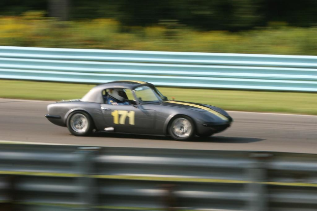 Ein Auto mit einem Helm tragenden Fahrer fährt auf einer Straße mit Absperrungen, mit Bäumen im Hintergrund.