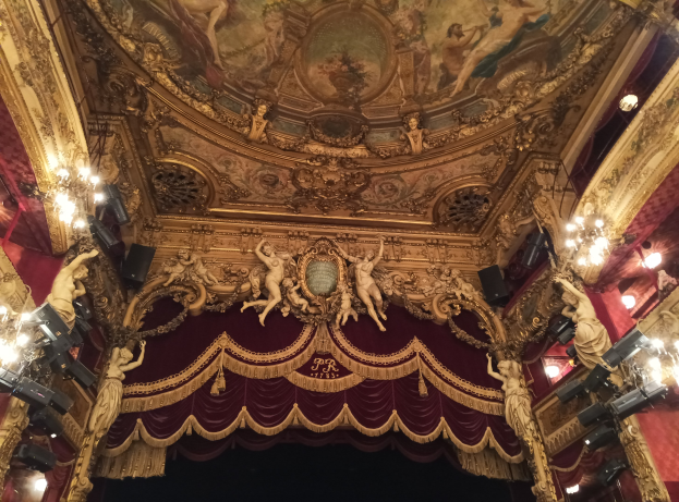 Theaterinnenraum mit roter Gardine, Kronleuchtern, Skulpturen, Lichtern und einer bemalten Decke, mit einer Bühne und einigen Menschen davor.