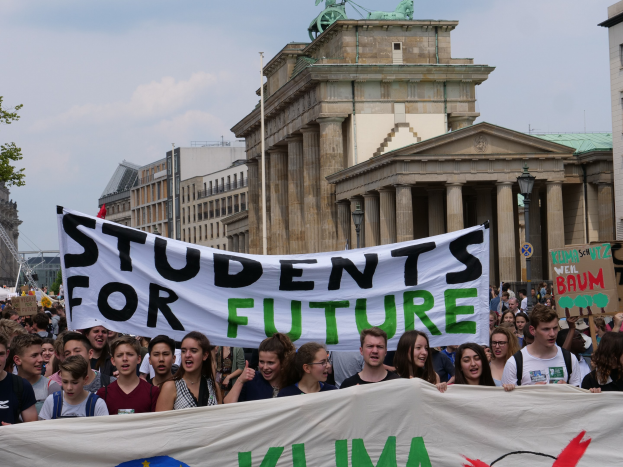 Schüler in Berlin marschieren mit buntem "Students for Future"-Schild vor Gebäude-, Baum- und Himmel-Hintergrund.