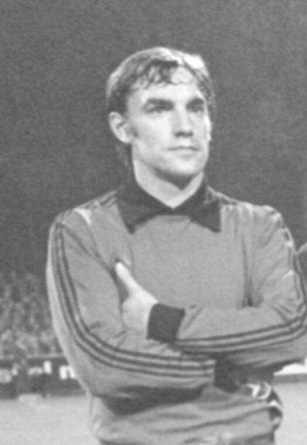 Schwarzes und weißes Foto eines Mannes, der mit verschränkten Armen auf einem Fußballfeld steht, umgeben von sitzenden Menschen im Hintergrund.