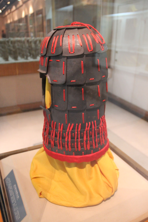 Helm in einer Glasvitrine mit einem gelben Tuch darüber, begleitet von einer Texttafel auf der linken Seite und Deckenleuchten im Hintergrund.