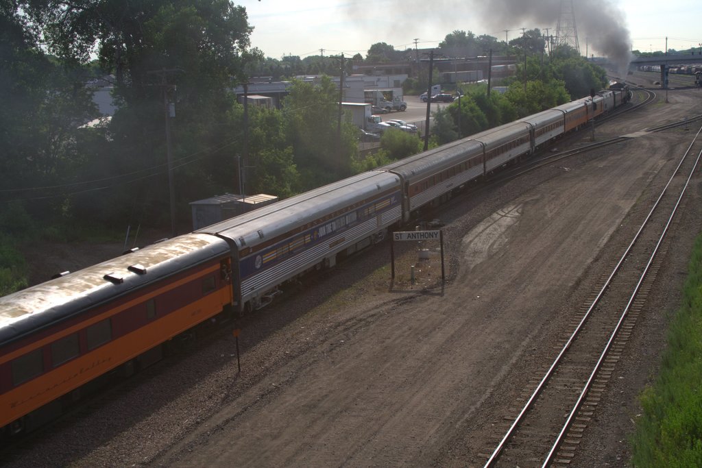 Ein Zug auf Schienen mit Bäumen, Strommasten mit Kabeln, Autos und Gebäuden links, eine Brücke vorne und Rauch, der aus dem Zug aufsteigt.