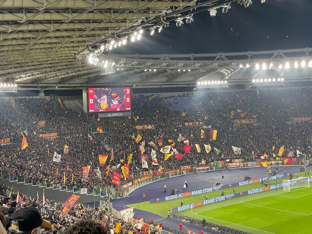 Eine große Menge in einem hell erleuchteten Stadion schwenkt Fahnen und Banner und wartet auf ein Fußballspiel, wobei einige Zuschauer Mützen tragen und ein Bildschirm im Hintergrund zu sehen ist.