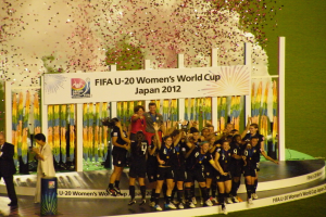 Frauenfußballspieler in schwarzer Uniform feiern auf einer Bühne mit Publikum und dekoriertem Hintergrund.