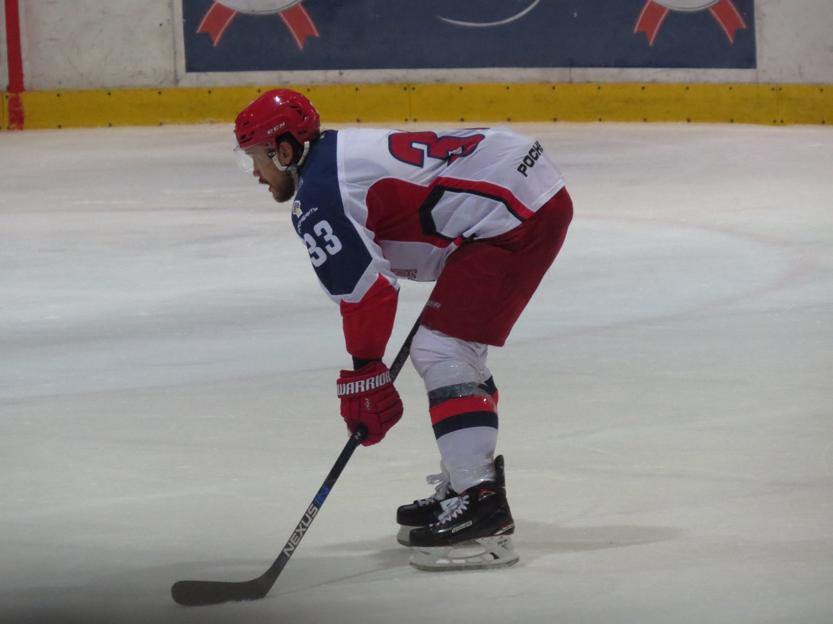 Hockeyspieler in voller Ausrüstung auf dem Eis mit einem Stock, der einer Wand mit Banner im Hintergrund gegenübersteht.