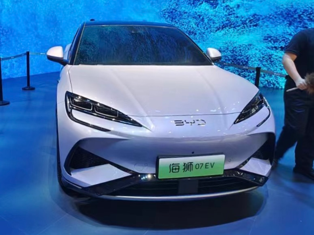 Ein weißes Auto auf einer Automesse, daneben steht eine Person mit einem Gegenstand in der Hand, im Hintergrund ein Bildschirm und auf der linken Seite Absperrböcke mit Seilen.