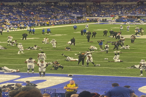 Ein Fußballspiel wird in einem Stadion mit Menschen auf dem Boden und einigen auf Stühlen im Hintergrund gespielt, während eine Person eine Kamera hält, was auf eine Live-Übertragung hinweist.