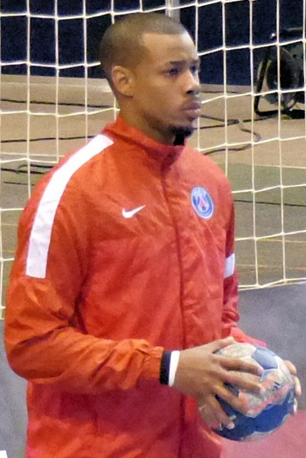 Ein Mann in einem blauen T-Shirt und Shorts steht vor einem Tor und hält einen Fußball, mit einem entschlossenen Gesichtsausdruck und konzentrierten Augen.