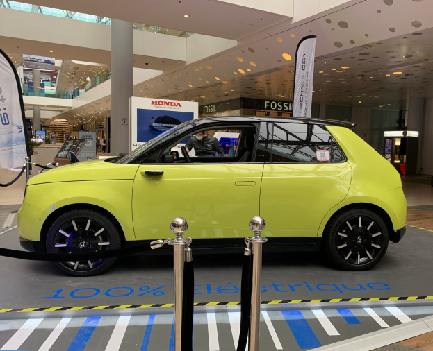 Gelber wasserstoffbetriebenes Elektroauto vor einem Gebäude mit Absperrpollern, Bannern, Menschen, Schildern, einer Topfpflanze, Deckenleuchten und Lüftern.