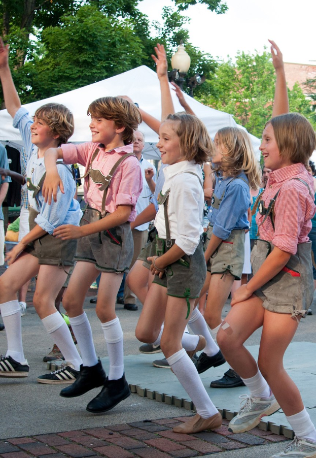 Eine Gruppe von Kindern in traditioneller bayerischer Tracht tanzt auf einer Straße, umgeben von einer Menge, Zelten, Laternenmasten, Bäumen, Gebäuden und einem klaren blauen Himmel, wobei einige Kinder Shorts und Schuhe tragen.