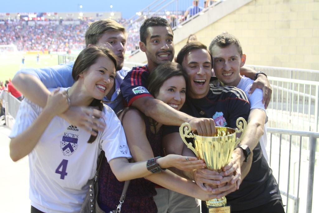 Sechs Menschen, zwei Frauen und vier Männer, halten einen Pokal und lächeln für ein Foto vor einem Eisenzaun, mit einer Wand und einem Zaun oben rechts, Menschen und Lichter im Hintergrund, möglicherweise in einem Stadion.