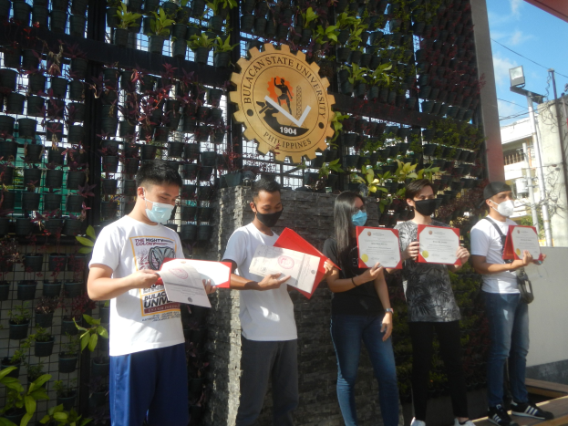 Eine Gruppe von Menschen in Masken hält Zertifikate und steht vor einem Universitätsgebäude mit einer bepflanzten Wand auf der linken Seite und einem gewölbten Eingang mit Logo und Text auf der rechten Seite.