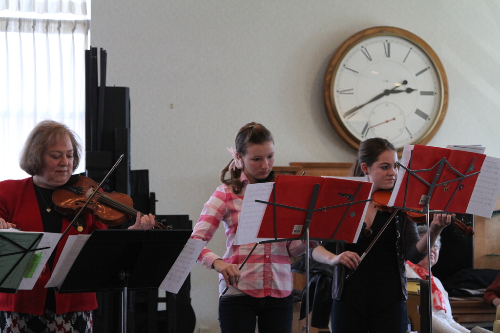 Drei Frauen stehen auf der Bühne und spielen Geigen, mit Notenpulten und Noten davor, während ein Zuschauer im Vordergrund sitzt; eine Uhr ist an der weißen Rückwand angebracht, und ein weißer Vorhang ist links sichtbar.