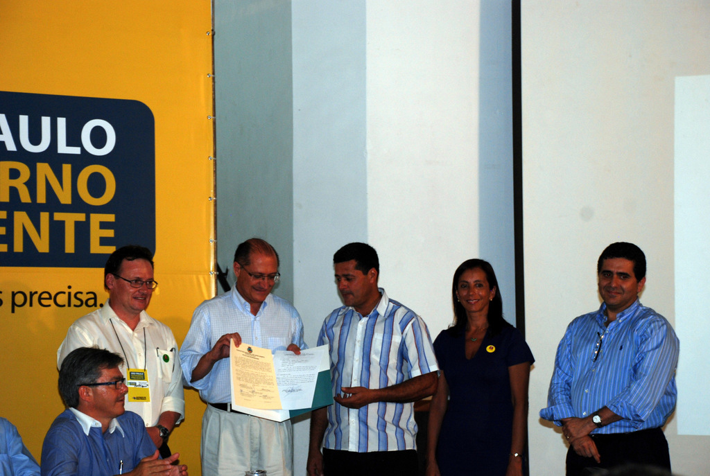 Eine Gruppe von Menschen, einige stehend und einige sitzend, mit einer Person, die eine Mappe mit Papieren hält, vor einer weißen Wand und einer gelben, weißen und blauen Fahne.