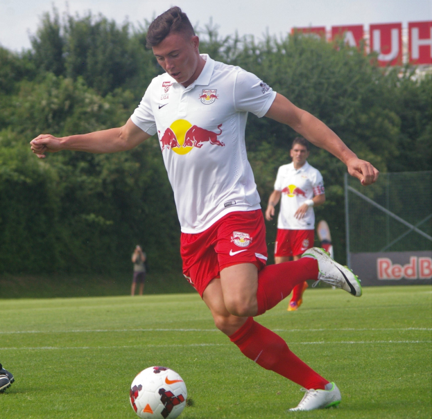 Ein professioneller Fußballspieler schießt einen Ball auf einem Rasenfeld mit Bäumen und einem klaren blauen Himmel im Hintergrund, wobei ein Schild mit der Aufschrift 'RB Leipzig' zu sehen ist.