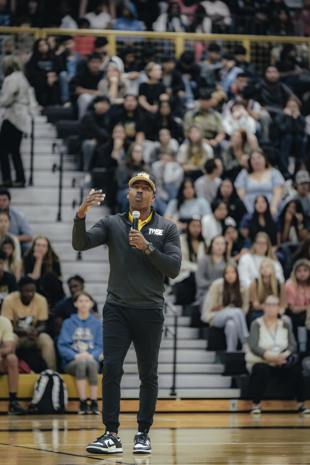 Ein Mann mit Mütze hält ein Mikrofon auf einem Basketballfeld, hinter ihm sitzen Menschen auf einer Treppe und Taschen liegen auf dem Boden.