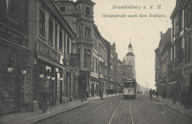 Ein Schwarz-Weiß-Foto einer Stadtstraße in Brandenburg, Deutschland, das eine Straßenbahn auf Schienen, Menschen auf dem Gehweg, Gebäude mit Fenstern, Schilder und Straßenlaternen unter einem bewölkten Himmel zeigt.