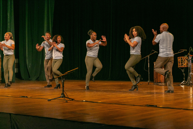 Gruppe von Menschen auf einer Bühne, die jubelnd klatschen, mit Mikrofonen, Musikinstrumenten und einem Vorhang im Hintergrund.