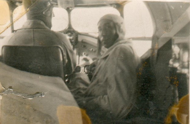 Zwei Männer in Schwarz-Weiß-Foto, einer sitzt im Cockpit in Pilotenuniform, der andere steht neben ihm.