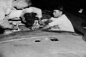 Drei Männer in Mützen arbeiten an einem schwarzen und weißen Auto in einer Garage, umgeben von Werkzeugen und Materialien.